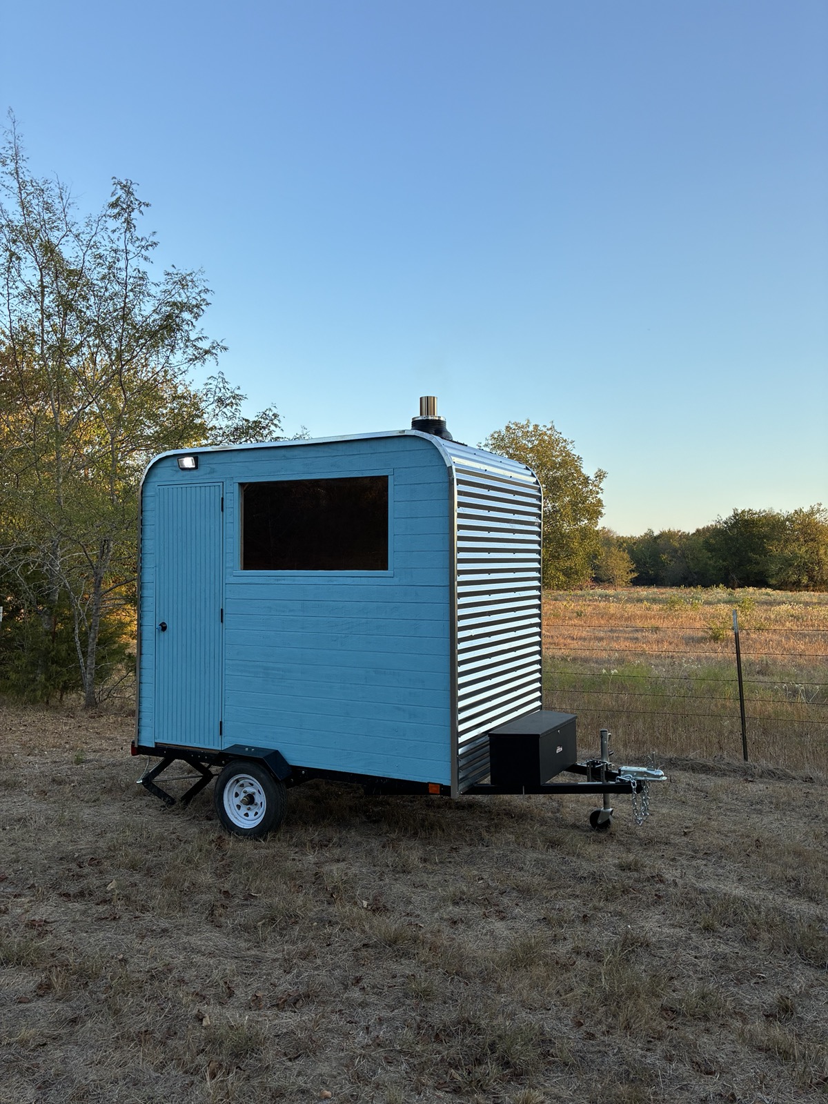 Sweatystoves trailer in daylight — sea-blue siding, corrugated steel, window, and hitch