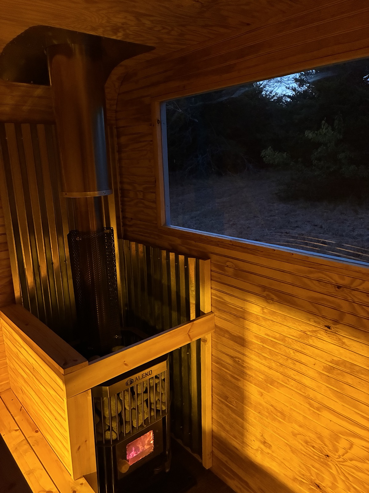 Wood-fired sauna stove with glowing fire, chimney, and a window looking out at dusk
